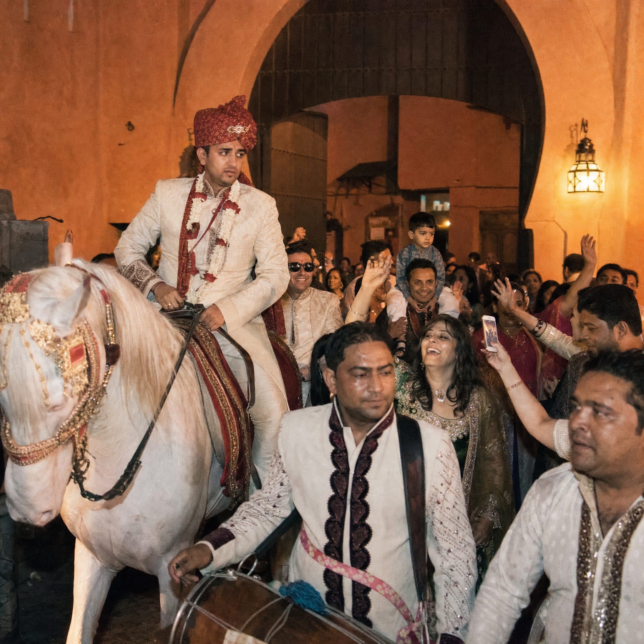 Groom arriving by white horse with dhol players during a Baraat at a palace gate