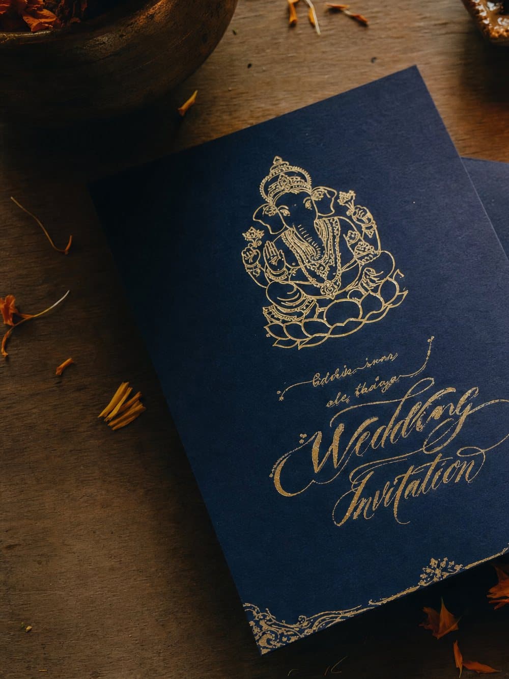 Navy and gold wedding invitation with gold calligraphy and marigold petals on a wooden table