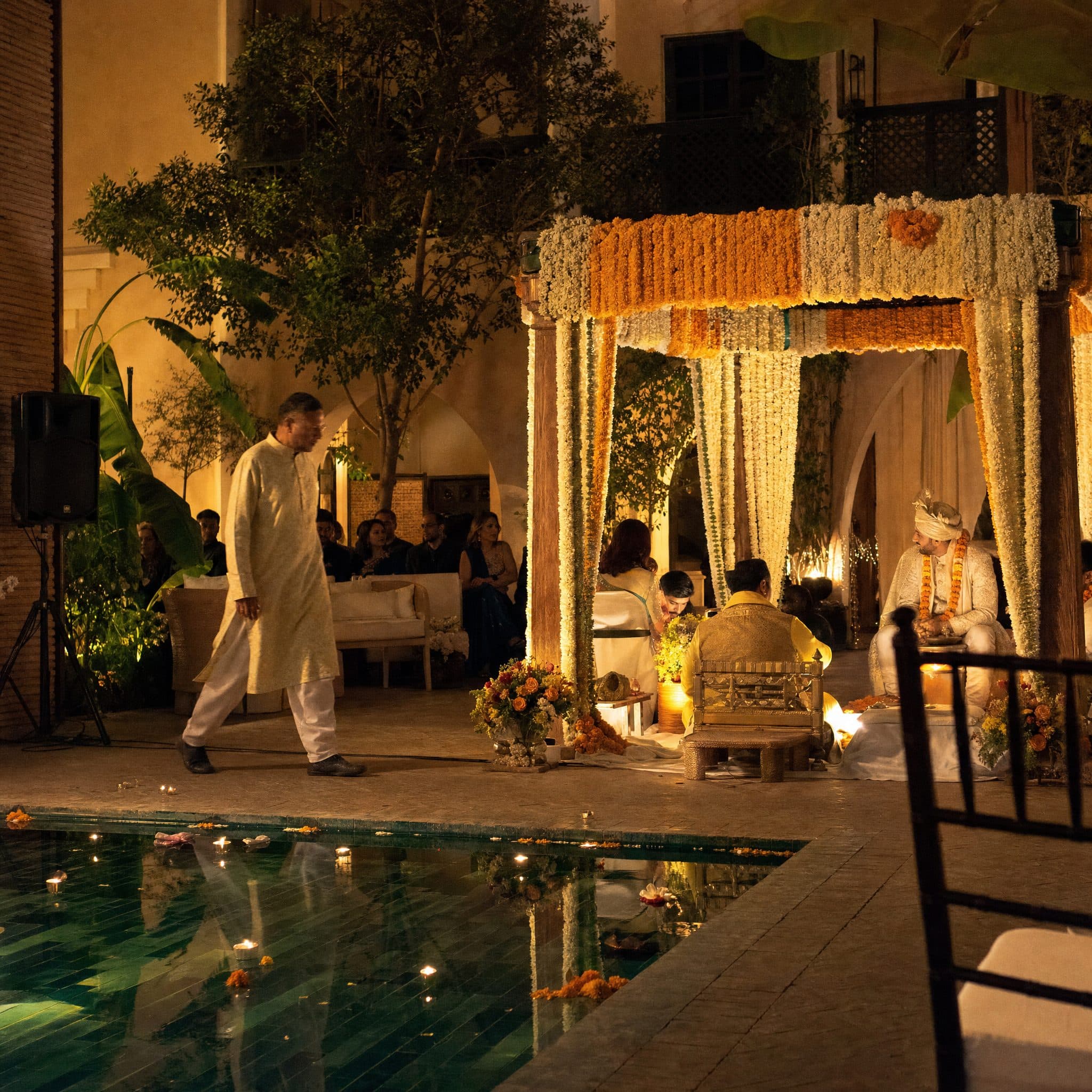 Mandap lit at night beside a reflecting pool with guests seated around it