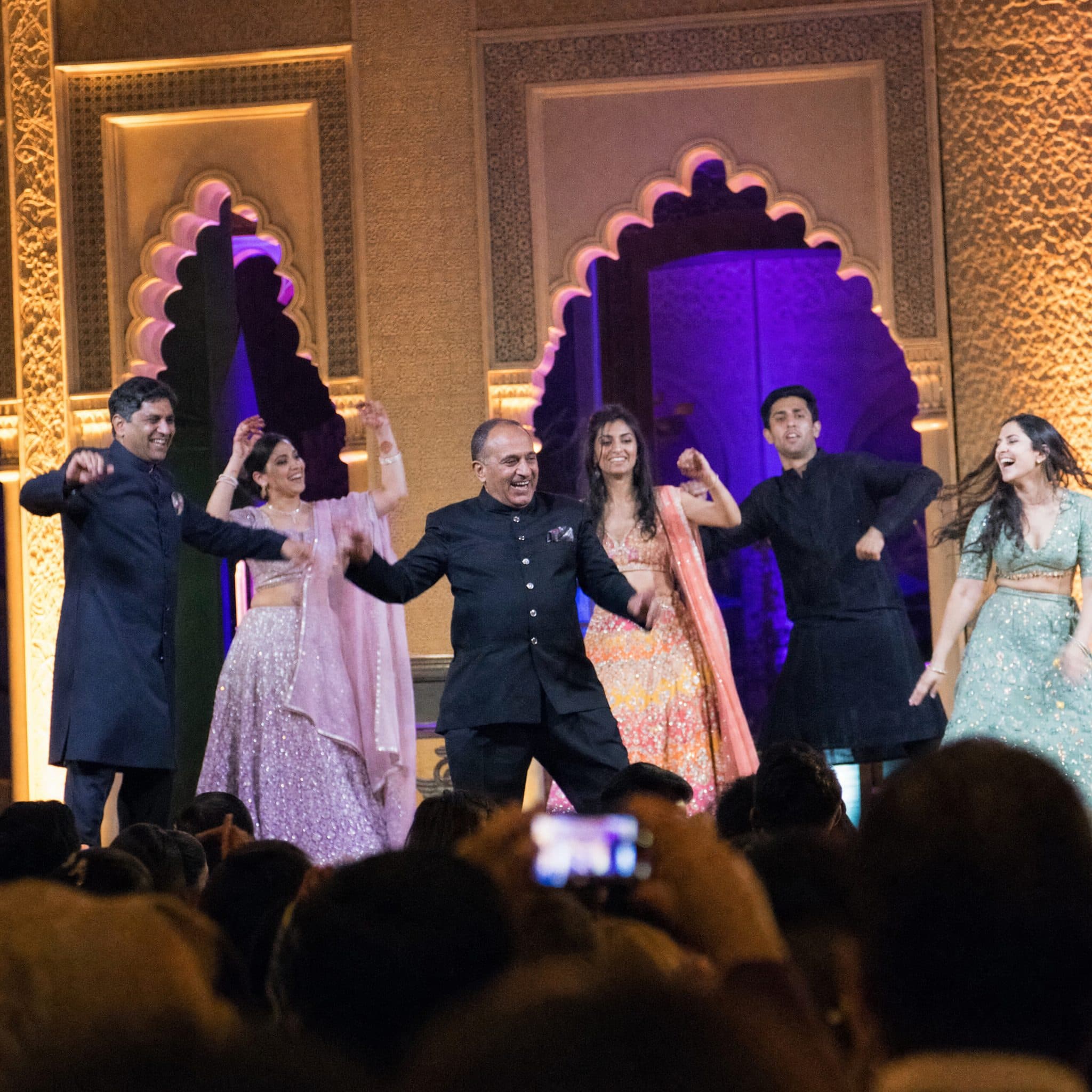 Family performing a choreographed Sangeet routine on a lit stage framed by Moorish arches